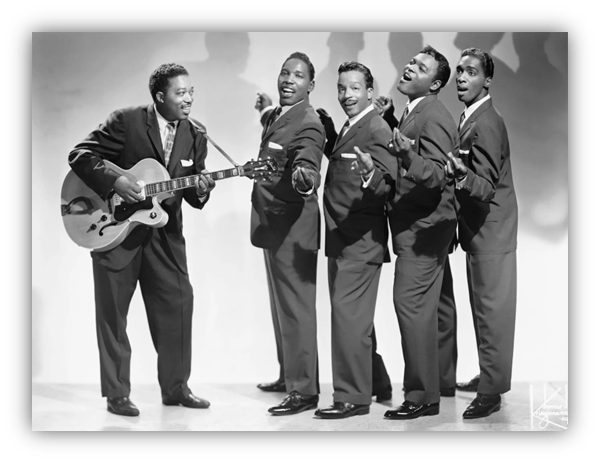 "Vintage black and white photo of five male musicians in suits, one playing guitar, performing joyfully."