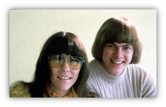 "1970s couple smiling, wearing sunglasses and casual sweaters."