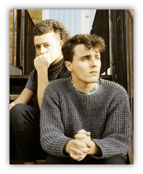 Two young men sitting on stairs, one with curly hair in a sweater, the other with a mohawk hairstyle.