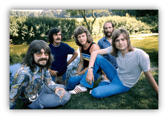 Five men sitting on grass by a serene river, dressed in 1970s fashion.
