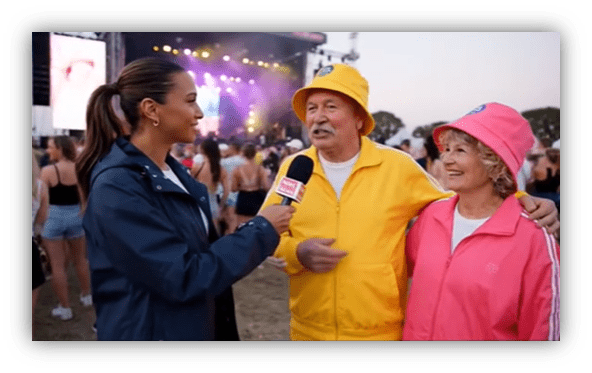 "Interview with a couple in colorful outfits at a music festival, with a performer on stage in the background."