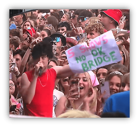 Crowd at a concert holding a sign that reads "5 Shows No OK Bridge" while an artist performs.