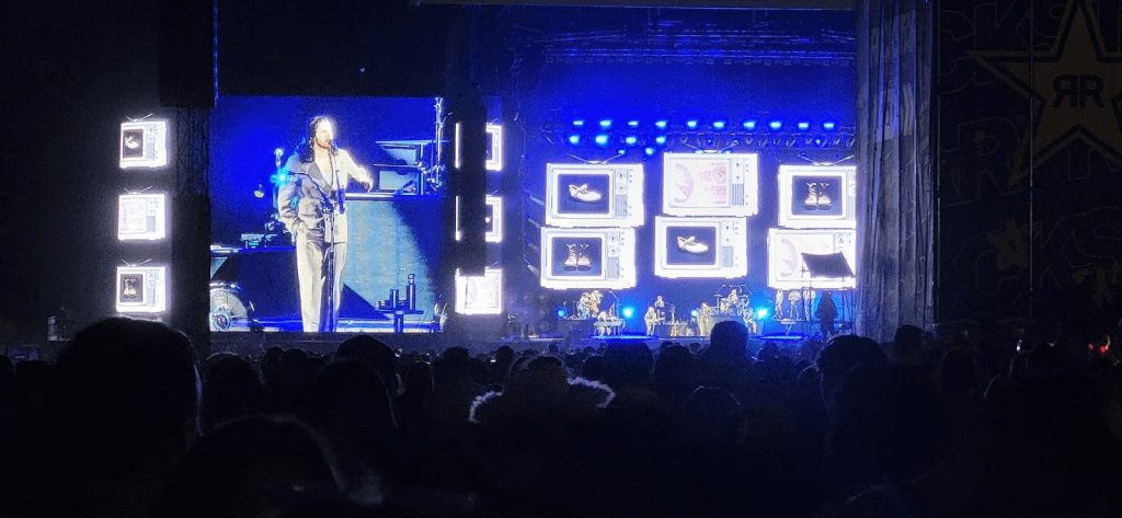 Concert performance with a singer on stage, illuminated by vibrant lights and screens displaying visuals.