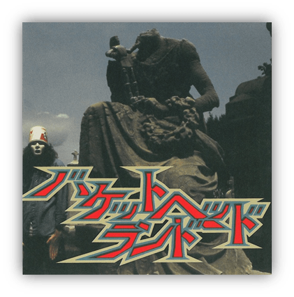 "Album cover featuring a statue and a person in a mask, with stylized text in Japanese."