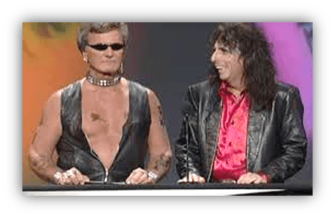 "Two male presenters in leather jackets on stage, one wearing sunglasses and a choker."