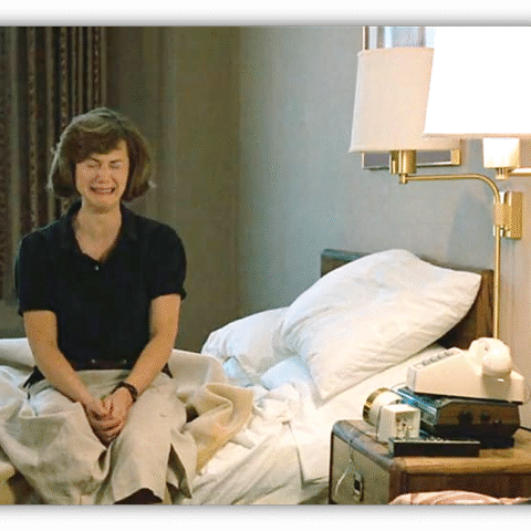 Woman sitting on a bed in a hotel room, expressing a mix of emotions.
