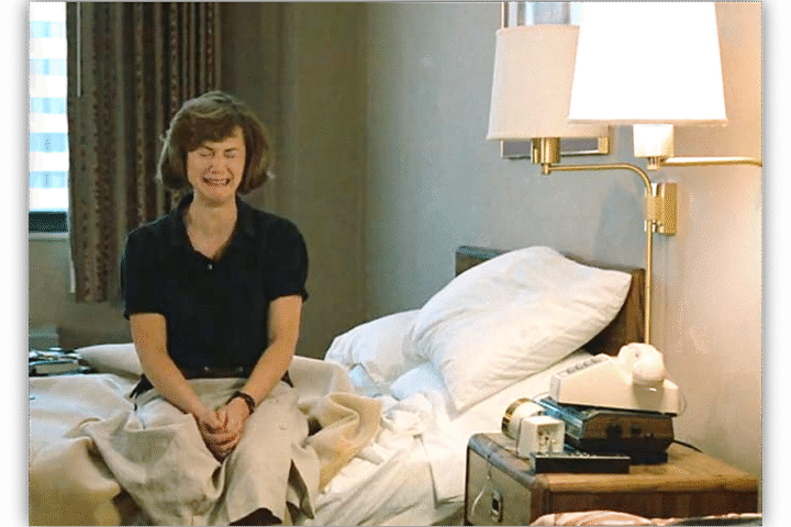 Woman sitting on a bed in a hotel room, expressing a mix of emotions.