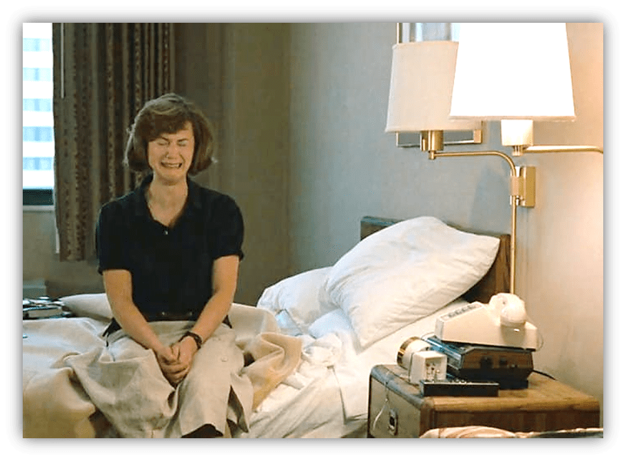Woman sitting on a bed in a hotel room, expressing a mix of emotions.