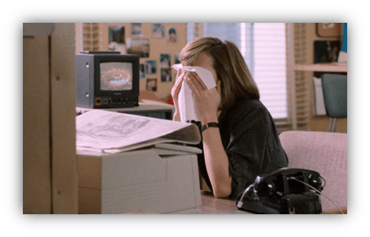 Woman sitting at a desk, covering her face with a tissue, looking distressed in an office setting.