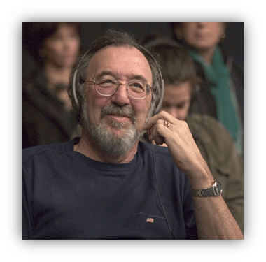 "Smiling man with glasses and headphones, seated in a crowd."