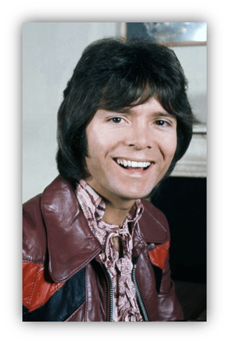 Smiling man with long dark hair wearing a leather jacket and patterned shirt.