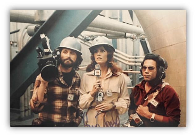 "Three journalists in safety helmets, one with a camera, capturing a story in an industrial setting."
