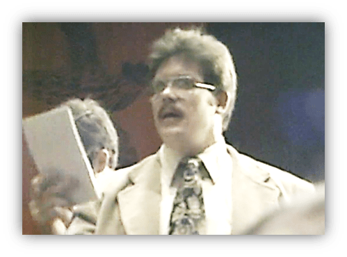 Man in a suit holding a paper, speaking at an event.
