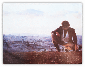 Man crouching on a hillside with a cityscape in the background.