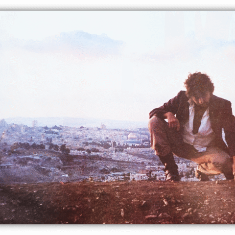 Man crouching on a hillside with a cityscape in the background.