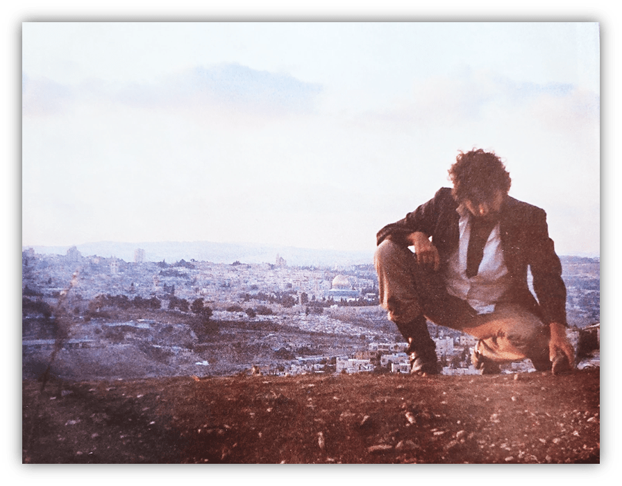 Man crouching on a hillside with a cityscape in the background.