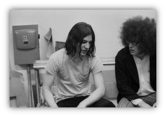 Two men engaged in conversation in a black and white photo, showcasing 1970s fashion and hairstyles.
