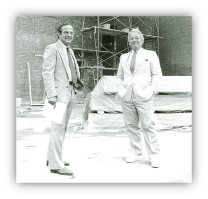 Two men in suits standing at a construction site, smiling.