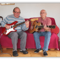 Two older men playing guitar together on a couch.