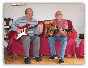 Two older men playing guitar together on a couch.