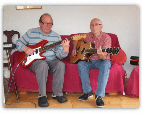 Two older men playing guitar together on a couch.