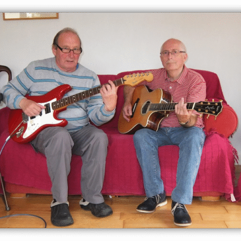 Two older men playing guitar together on a couch.