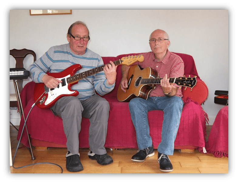Two older men playing guitar together on a couch.