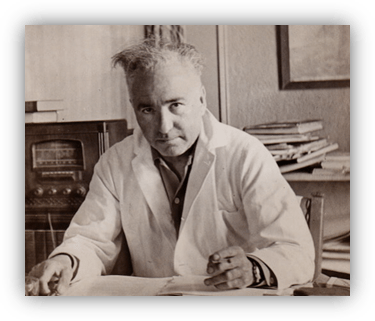 Black and white photo of a scientist in a lab coat, seated at a desk with papers and books.