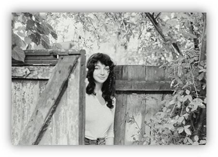 "Black and white photo of a woman with curly hair standing by a wooden gate surrounded by greenery."