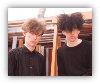 Two young men with unique hairstyles, one in a black shirt and the other in a black long-sleeve top, standing against a backdrop of wooden structures.