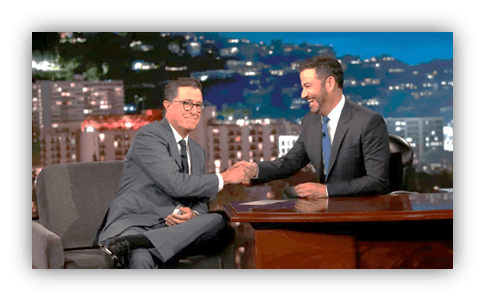 "Stephen Colbert and Jimmy Kimmel shaking hands on late-night talk show set."