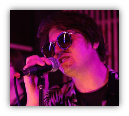"Musician singing into a microphone, wearing sunglasses and a patterned shirt, under purple stage lights."