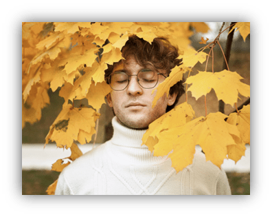 Young man with glasses standing among vibrant yellow autumn leaves, eyes closed, wearing a cozy white turtleneck sweater.
