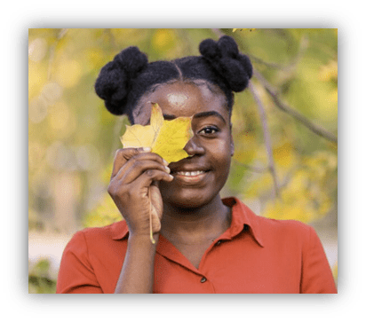 Smiling woman holding a yellow leaf in front of her face, surrounded by autumn foliage.