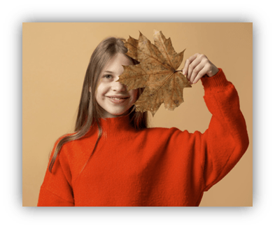 Smiling girl in an orange sweater holding a large autumn leaf against a beige background.