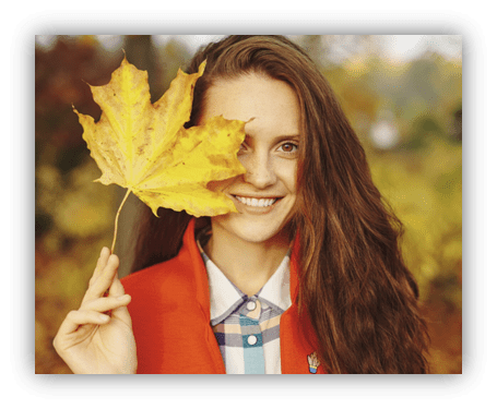 Smiling woman holding a yellow maple leaf in autumn scenery.