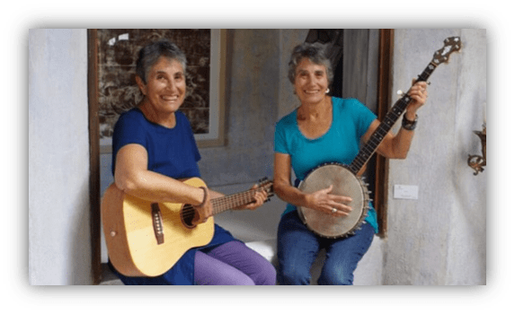 Two women playing guitar and banjo indoors, smiling and enjoying music together.