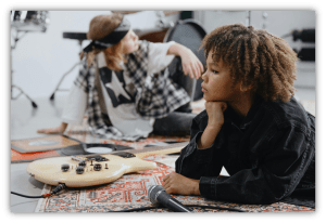 Two children in a music studio, one laying with a guitar and the other seated, both engaged in creative expression.