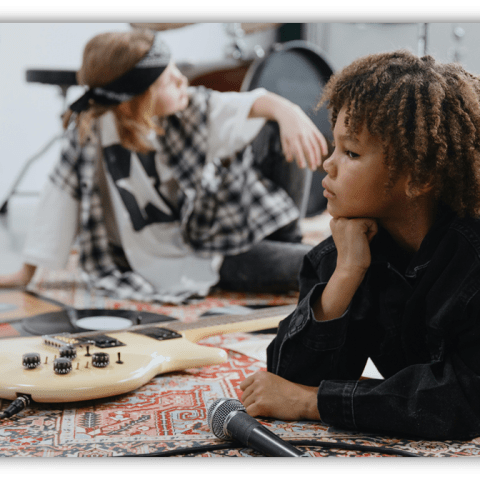 Two children in a music studio, one laying with a guitar and the other seated, both engaged in creative expression.
