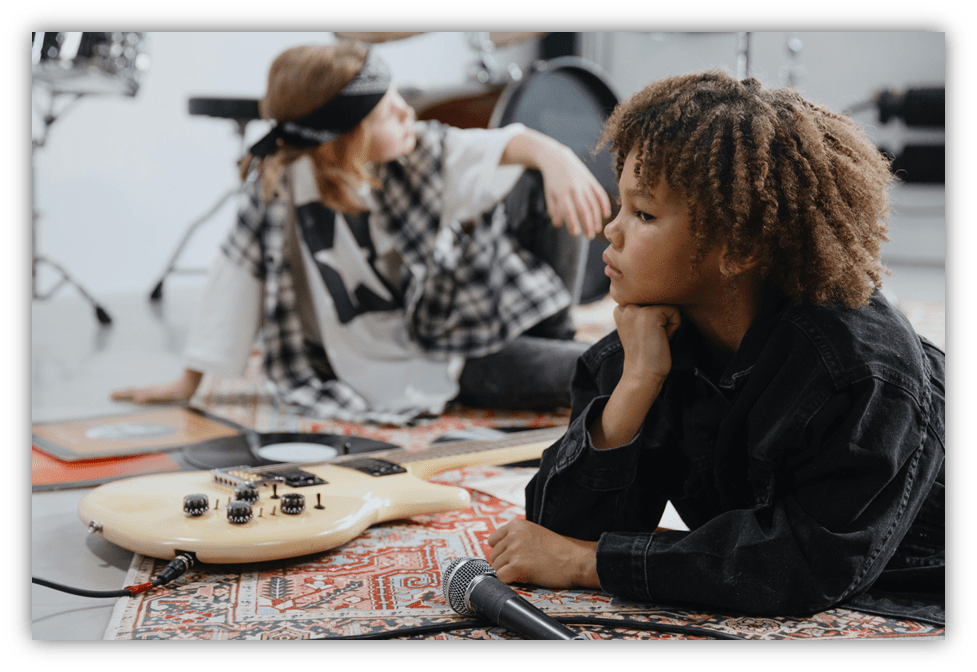 Two children in a music studio, one laying with a guitar and the other seated, both engaged in creative expression.