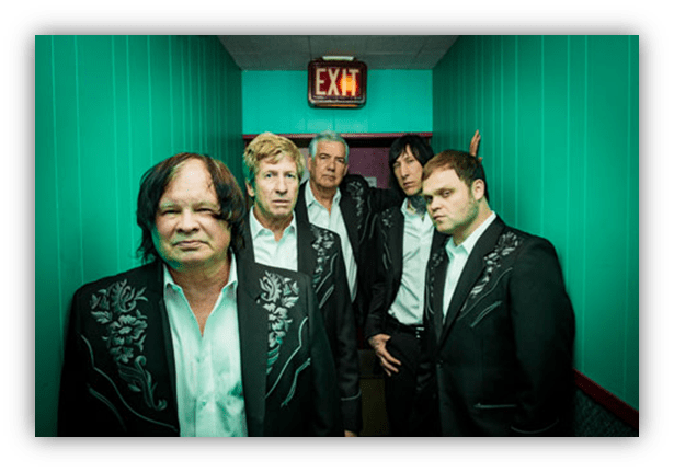 "Five musicians in black suits with floral designs posing in a turquoise hallway."