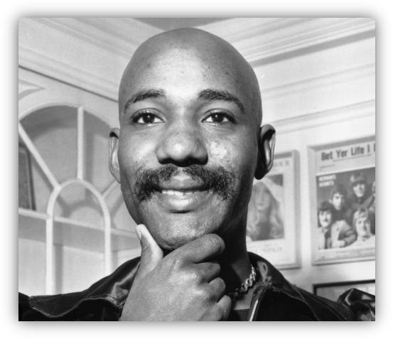 Black and white portrait of a smiling man with a mustache, wearing a leather jacket and posing thoughtfully.