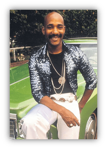 "Man in sequined jacket posing beside a classic green car."