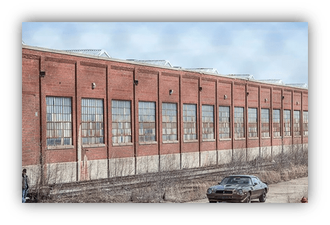 "Brick warehouse exterior with large windows and vintage car parked nearby."