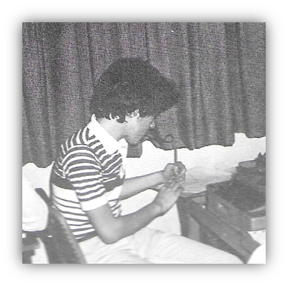 "Young man writing at a desk with a typewriter, wearing a striped shirt."