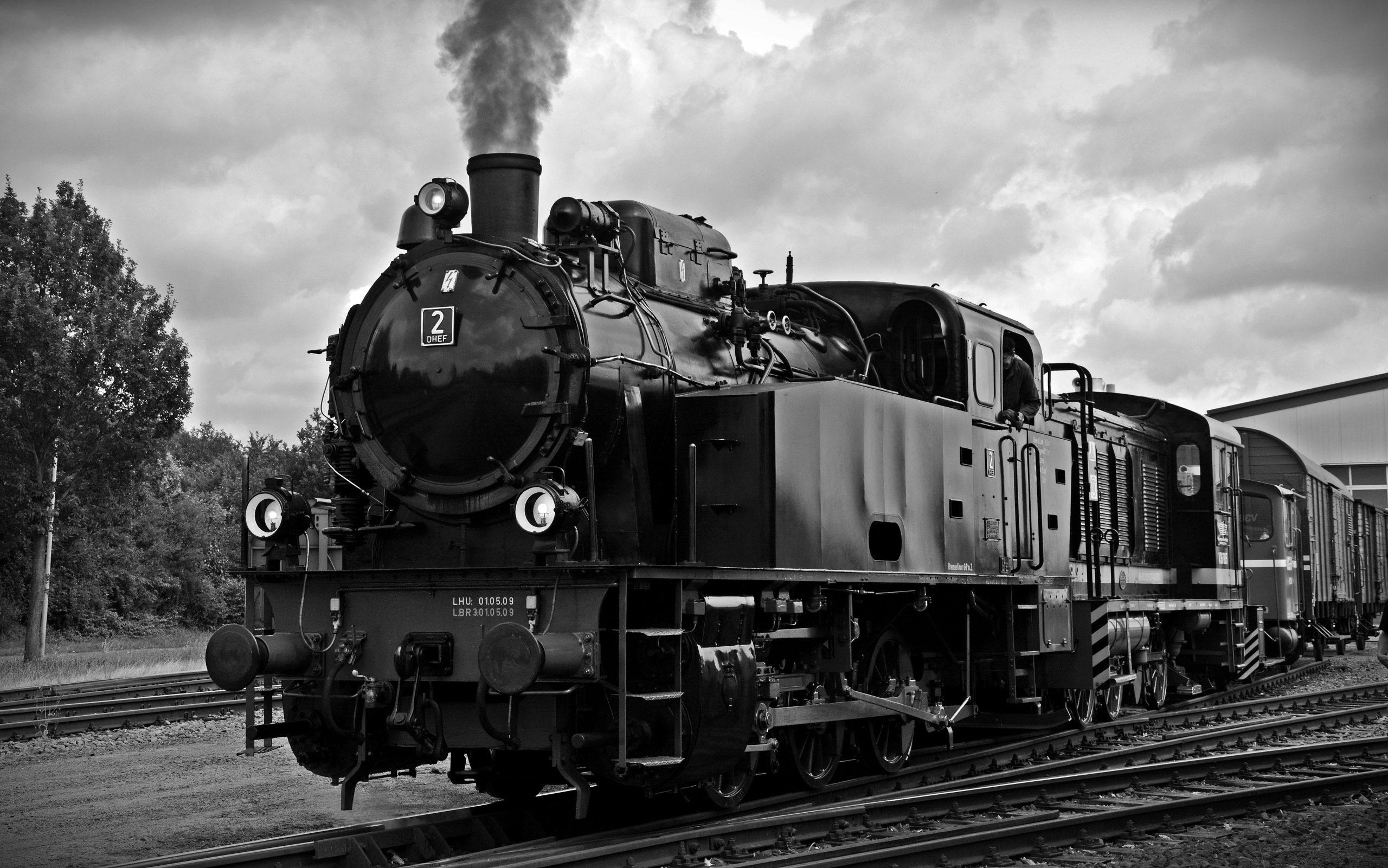 Black and white image of a vintage steam locomotive with smoke billowing, parked on train tracks.